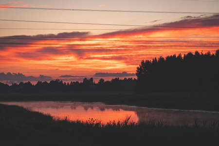 sunset over the river. Red orange clouds. Black forest silhouette over the river. Electric power line wires in he air. Reflection of clouds and reeds in river makes it orangeの写真素材