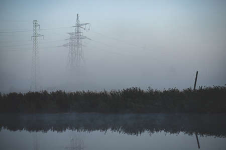 A power line passes over the river in Latvia. Morning shot in the haze on the background of the forest. Lots of pylons in meadow looks like big giantsの写真素材