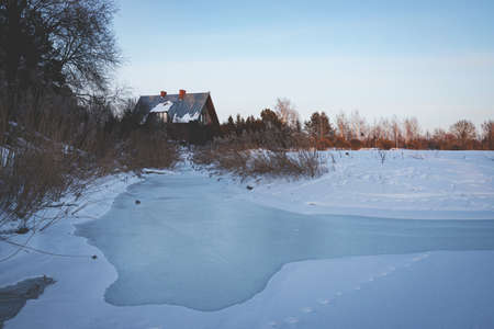 ice and snow covered water, big countryside house between the trees, dry yellow river reeds, blue clear sky, winter evening sunset light, usual landscape in Latviaの写真素材