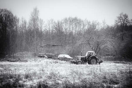 old soviet tractor and wreck of small automobile in snow covered field near abandoned house in Latviaの写真素材
