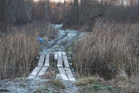 dangerous unstable hand made bridge over small river in Latvia. First snow on ground, forest ahead, blue barral left on road side, hard dirt road for 4x4 vehiclesの写真素材