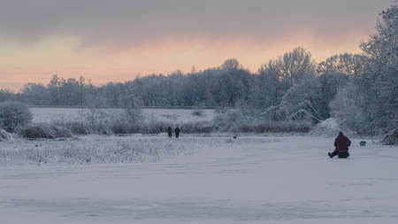 three fishermen on ice and snow covered lake near road, all ground and trees covered with frost and...