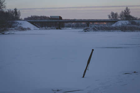 river scene in wintertime, snow and ice covered river, transport bridge with heavy truck in distance, ice breaking chisel for making holes for fishing on ice, December evening in Latvia, river Vircavaの写真素材