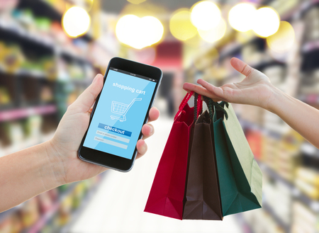 hand holding mobile smart phone with mobile shop  on supermarket blur background and shopping bags - e-commerce concept