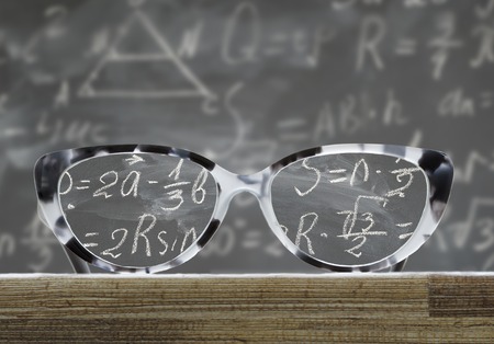 glasses on a wooden table in front of blackboard with math formulas, educational back to school conceptの写真素材