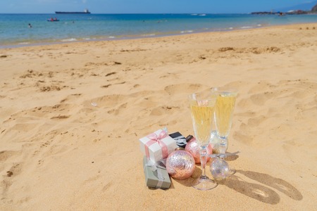 Two glasses of christmas champagne with bubbles and christmas gift boxes and decorations on beach by the tropical seaの写真素材