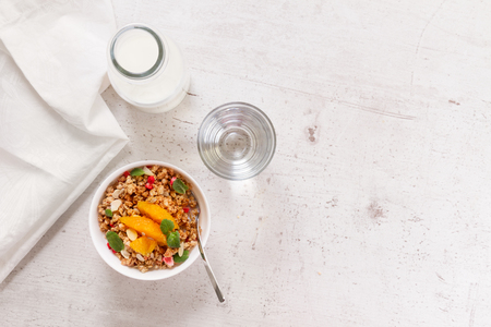 Healthy breakfast - plate of granola with orange slices, mint and seeds, bottle of milk, water, copy space on white wooden tableの写真素材