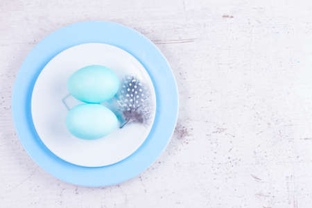 Plates with two painted blue eggs and feather on white wooden tabletopの写真素材