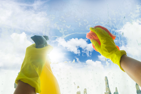 Spring cleaning - someones hands in yellow gloves with spray and ruber cleaning window, spring blue sky in backgroundの写真素材