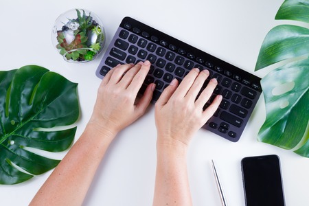 Flat lay home office workspace with someones hands typing on black keyboard and green leaves on white backgroundの写真素材