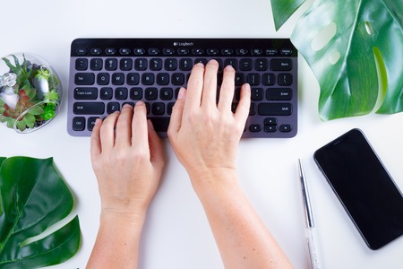 Flat lay home office workspace with someones hands typing on black modern keyboard and green leaves on white backgroundの写真素材