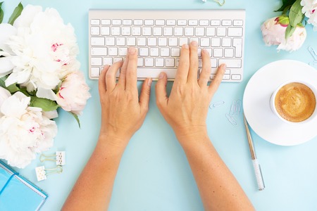Flat lay home office workspace frame with someones hand typing white modern keyboard, notebook and peony flowers on blue backgroundの写真素材