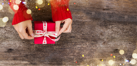 Christmas gift giving - someones hand in red knitted sweater making bow on red box with present, banner with copy spaceの写真素材