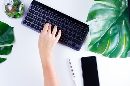 Flat lay home office workspace with someones hands typing on black modern keyboard and green monstera leaves on white backgroundの写真素材