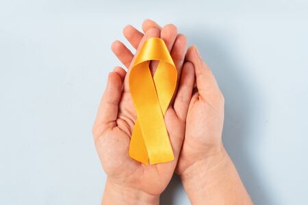 Two hands holding Childhood Cancer Awareness Yellow Ribbon on blue background with copy space. Childhood Cancer Day February, 15の写真素材