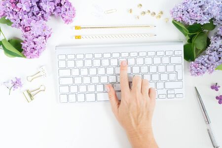 Flat lay top view home office workspace - hand typing on modern keyboard with lilac flowers on white