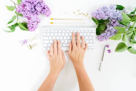 Flat lay top view home office workspace - two hands typing on modern keyboard with lilac flowers on white backgroundの写真素材