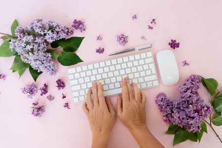 Flat lay home office workspace - modern keyboard with two hands typing, fresh lilac flowers