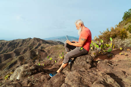 Senior man with laptop on the edge of a mountain with stunning views of the valley, healthy...