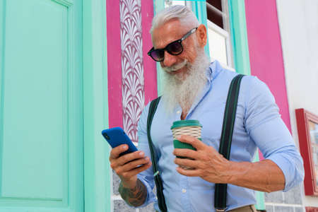 Senior hipster man in black glasses having fun with a modern phone on a europen city street, close...