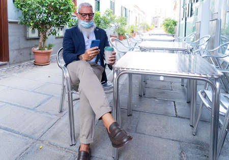Senior man in mask with modern phone in cafe during coronavirus outbreak, new normal elderly life conceptの写真素材