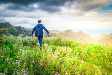 Rear view of young man hiker traveler standing on dirt mountain path in nature reserve on green prairie scenery in cloudy weather in warm jacket. Travel, adventure, hiking and motivation conceptの写真素材