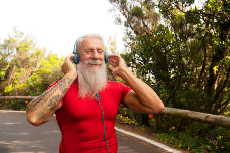 Senior athlete man with white beard doing running outdoor on the nature, listening music in...