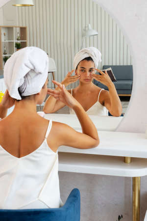 Skin care. Beautiful young brunette woman looking at herself doing face fitness - self face massage,...