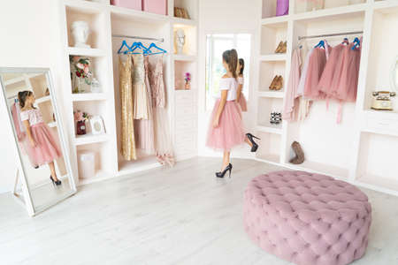 Rear view of tender teen girl in pink tutu-skirt and black high-heeled shoes posing in front of mirror in wardrobe room, looking at own reflectionの写真素材
