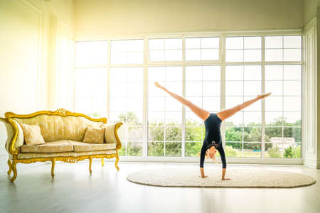 Teenage girl gymnast in sport bodysuit makes gymnastics and stretching routine by raising legs up and standing on hands in room with big windowsの写真素材