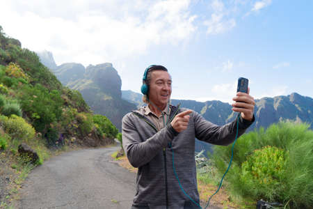 Happy middle aged man traveler smiling at phone against of hill, blue sky and beautiful landscape,...