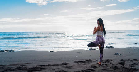 Life balance. Full length of slim sporty woman in sports wear doing tree pose or vrikshasana while practicing yoga and meditation on seashore in peaceful morning, rear view. Healthy lifestyle conceptの写真素材