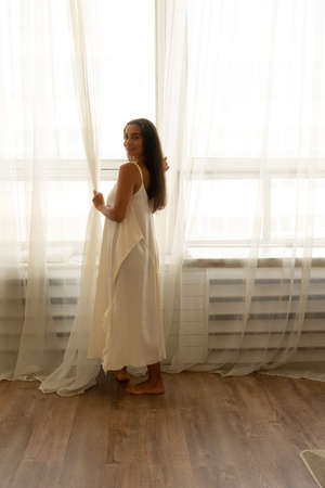 New day. Rear view of young brunette woman after waking up stands by large window in silk white gown and opening curtains with both hands, letting first morning rays of sun into bedroom at homeの写真素材