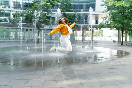 Happy curvy woman in city, plus size brunette lady with curly gray hair jumping emotionally in front of the fountains in modern cityの写真素材