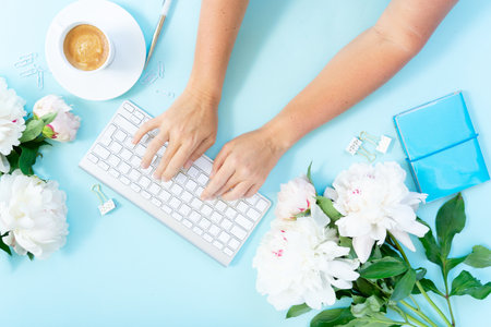 Flat lay home top view office workspace frame with someones hand typing white modern keyboard, notebook and peony flowers on blue backgroundの写真素材