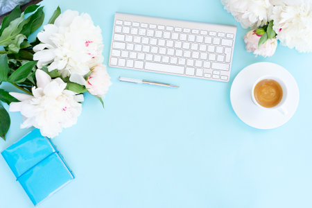 Flat lay home office workspace frame with white modern keyboard, notebook and peony flowers, copy space on blue backgroundの写真素材