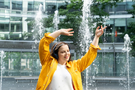 Happy curvy woman in city, plus size brunette lady with curly hair dancing in headphones emotionally in front of the fountains in modern cityの写真素材