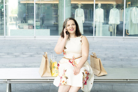 Happy curvy woman with ecological shopping bags sitting and talking on mobile phone enjoying in shopping. Consumerism, shopping, lifestyle conceptの写真素材