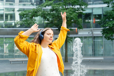 Happy curvy woman in city, plus size brunette lady with curly hair dancing in headphones emotionally in front of the fountains in modern cityの写真素材