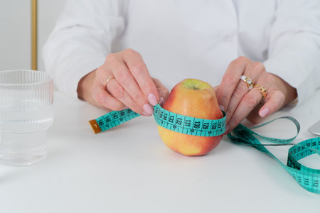 A smiling woman in a white shirt presents an apple wrapped in a measuring tape, symbolizing healthy...
