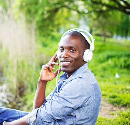 Young happy smiling african american sitting on a grass and listens to music in a parkの写真素材