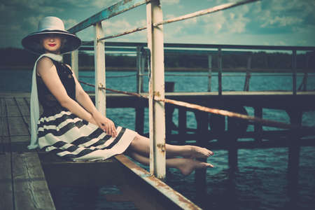 Beautiful woman wearing hat and white scarf sitting on old wooden pier の写真素材