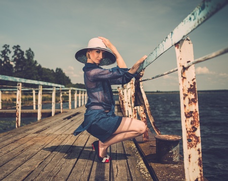 Stylish woman in white hat sitting near rails of old wooden pierの写真素材