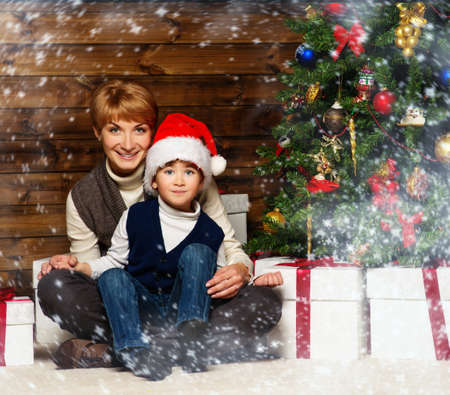 Mother and her lIttle boy in Santa hat with gift box under christmas tree in wooden house interior の写真素材