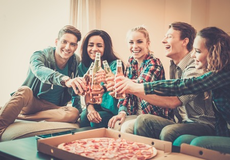 Group of young multi-ethnic friends with pizza and bottles of drink celebrating in home interior