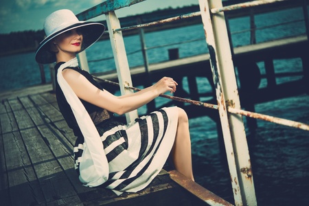 Beautiful woman wearing hat and white scarf sitting on old wooden pierの写真素材
