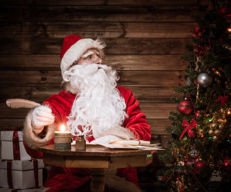 Santa Claus in wooden home interior sitting behind table and writing letters with quill penの写真素材