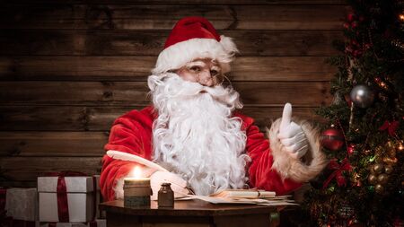 Santa Claus in wooden home interior sitting behind table and writing letters with quill penの写真素材