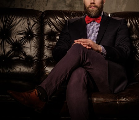 Old-fashioned bearded man sitting in comfortable leather sofa isolated on gray.の写真素材