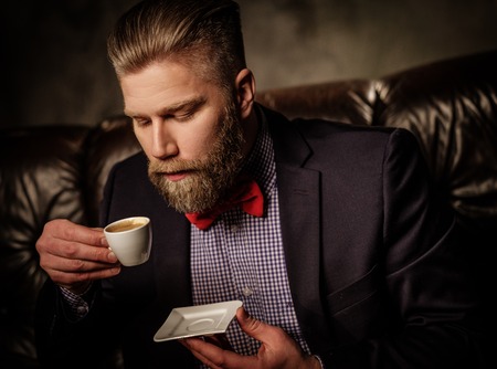 Old-fashioned bearded man sitting in comfortable leather sofa with cup of coffee isolated on gray.の写真素材